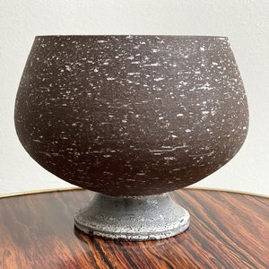 Puede incluir: Un cuenco grande de cerámica marrón con un acabado moteado se encuentra sobre una mesa de madera. El cuenco tiene una base ancha y redondeada y un borde acampanado.