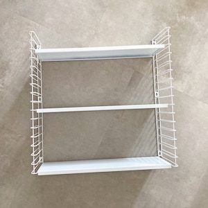 May include: White, three-tiered wall-mounted shelf. The shelves are rectangular and supported by a white wireframe. The design is minimalist and modern, suitable for displaying items or storage. The shelf is mounted on a neutral-coloured wall.