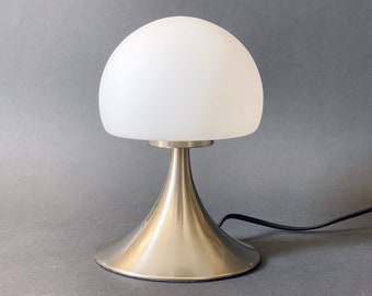 May include: A modern table lamp with a white, dome-shaped glass shade. The lamp has a brushed metal base and stem, and a black power cord. The design is minimalist and functional, suitable for a desk or bedside table.