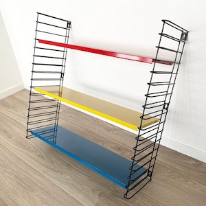 May include: A wall-mounted shelving unit with three shelves in red, yellow, and blue. The shelves are supported by a black wire frame. The unit is designed for displaying items or storage.