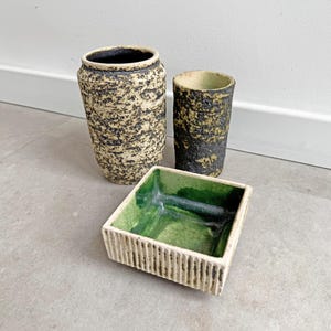Puede incluir: Tres piezas de cerámica: un jarrón alto, un vaso más corto y un plato cuadrado. El jarrón y el vaso tienen un diseño texturizado y moteado en beige y negro. El plato es blanco con interior verde.