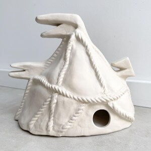 Könnte beinhalten: Eine matte, cremeweiße Keramikskulptur in Form eines Zeltes oder einer Hütte mit erhabenen, seilartigen Texturen, die in einem Gittermuster über die Oberfläche verlaufen. Die Struktur weist drei hervorstehende spitze Elemente sowie eine kreisförmige Öffnung auf.