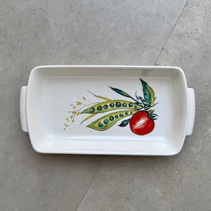 Peut inclure: Plat rectangulaire en céramique blanche avec un motif de légumes peint. Le motif présente des gousses de pois vertes, une tomate rouge et des feuilles vertes. Le plat a deux poignées de chaque côté.