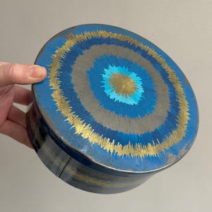May include: A round, blue decorative tin with concentric circles in shades of blue, grey, and gold. The lid and sides are painted with a radial design. The tin is held by a hand.