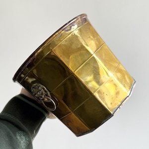 Può includere: Un secchiello per il ghiaccio vintage in ottone con un motivo geometrico. Il secchiello ha una finitura dorata, un bordo argentato e una maniglia a testa di leone. Il secchiello è tenuto da una persona.