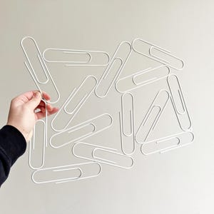 以下が含まれることがあります： 複数の白いワイヤークリップを幾何学的に重ね合わせて連結した、大型の白いペーパークリップ型彫刻。軽量でオープンフレーム構造の装飾的なウォールアート作品として構成されています。