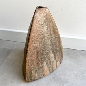 May include: A tall, triangular vase with a textured, weathered appearance. The vase has a brown and tan color palette with hints of green, and a rectangular opening at the top. The vase is likely made of ceramic or stoneware.