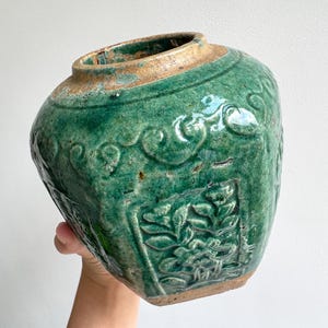 Peut inclure: Un pot en céramique hexagonal avec une glaçure vert émeraude brillante. Le récipient présente des motifs floraux en relief sur chaque face et une large ouverture circulaire avec un bord beige non émaillé et texturé.