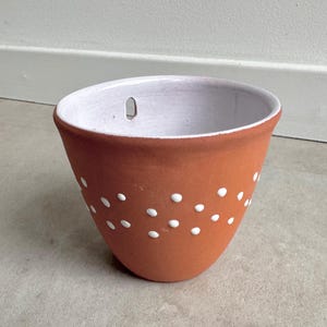 May include: A terracotta-colored ceramic planter with a white interior and a series of white polka dots. The planter has a rounded shape and a hole for hanging. A decorative item for indoor or outdoor use.