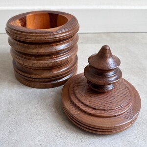Pu&ograve; includere: Una scatola di legno con un coperchio. La scatola &egrave; in legno scuro e ha una forma arrotondata. Il coperchio &egrave; anch'esso in legno e ha un pomo decorativo in cima. La scatola &egrave; vuota.