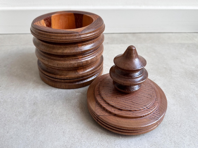 Pu&ograve; includere: Una scatola di legno con un coperchio. La scatola &egrave; in legno scuro e ha una forma arrotondata. Il coperchio &egrave; anch'esso in legno e ha un pomo decorativo in cima. La scatola &egrave; vuota.