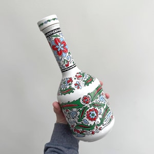 Op de afbeelding: Witte keramische fles met een lange hals en een afgeronde basis. De fles is versierd met een bloemmotief in rood, blauw en groen. De fles wordt door een persoon vastgehouden.