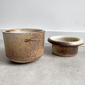 Könnte beinhalten: Zwei Keramik-Pflanzgefäße mit einer strukturierten, erdigen Optik. Der größere Pflanztopf ist zylindrisch, der kleinere hat einen abgerundeten, ausgestellten Rand. Beide haben eine braune und beige Farbpalette, die eine natürliche, rustikale Ästhetik suggeriert.