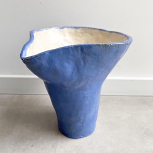 May include: A handcrafted, blue ceramic vase with an unusual, organic form. The vase features a broad, irregular rim and a textured finish. The interior is an off-white colour. This vase is a decorative item for the home.