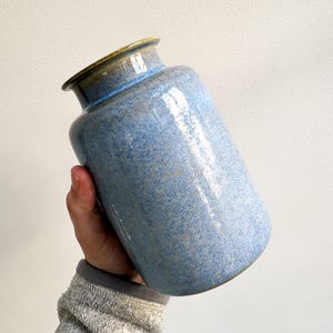 May include: A blue ceramic vase with a speckled texture and a glossy finish. The vase has a cylindrical body and a slightly flared rim with a brown and blue accent. The vase is held at an angle.