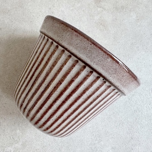 Może przedstawiać: Biała ceramiczna doniczka z brązowymi pionowymi paskami. Doniczka ma zaokrąglone dno i szeroki, rozszerzający się wierzch.