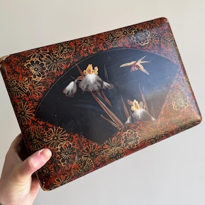 Può includere: Scatola antica rettangolare con sfondo nero con un disegno a ventaglio con fiori bianchi e gialli e un uccello in volo. La scatola ha un motivo floreale rosso e oro sui bordi.