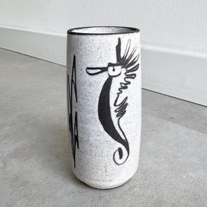 May include: A cylindrical ceramic vase with a speckled white finish. A black seahorse illustration is on the front, with the word "AQUA" in black vertical lettering. The vase has a dark rim and is suitable for home decor.