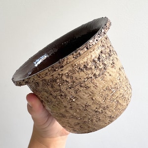 Op de afbeelding: Een cilindervormige keramische plantenpot met een ruwe, getextureerde buitenkant in tinten bruin en beige. De pot heeft een brede, uitlopende rand en een gladde, donkerbruin geglazuurde binnenkant. Het buitenoppervlak heeft een tactiel, ruw uiterlijk met donkerbruine spikkels over het lichtbeige kleilichaam.