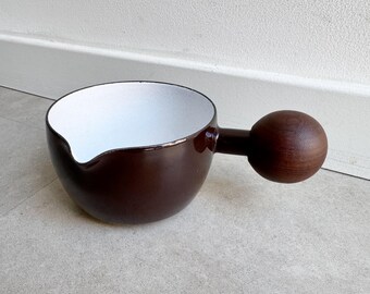 May include: A dark brown ceramic sauce pan with a smooth, glossy exterior and a matte white interior. The vessel features a small pouring spout on the rim and a distinct, large, spherical wooden handle extending from the side of the bowl.