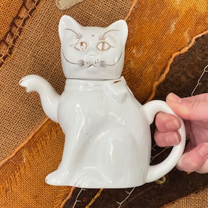 Op de afbeelding: Een witte keramische theepot in de vorm van een kat met gouden accenten op het gezicht en een kleine strik. De theepot heeft een handvat en een tuit, en de deksel is de kattenkop. Op een bruin en oranje getextureerd oppervlak.