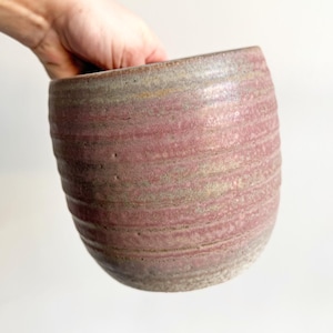 May include: A handmade ceramic vase with a rounded shape and a textured, ridged surface. The vase features a blend of colors, including shades of pink, gray, and beige. The vase is held by a hand, showcasing its size and form.