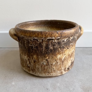 Pode incluir: Um vaso de cerâmica artesanal com uma aparência rústica e texturizada. O vaso tem uma forma cilíndrica com duas pequenas alças e uma abertura larga. A paleta de cores inclui tons de castanho e bege, criando uma estética natural e terrosa.