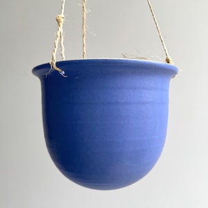 Può includere: Un vaso sospeso in ceramica blu, di forma arrotondata e con un bordo leggermente svasato. Il vaso è sospeso da una corda in fibra naturale. Il colore blu è uniforme e la superficie appare liscia.