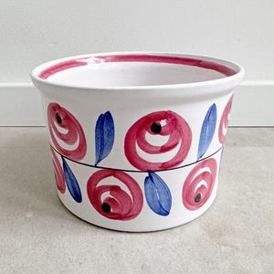 Pode incluir: Vaso de cerâmica branco com um design floral pintado à mão. O vaso apresenta rosas vermelhas, folhas azuis e uma borda vermelha. Uma linha preta separa as duas fileiras de flores. Decorativo, adequado para uso interno ou externo.