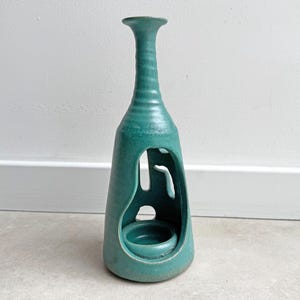 Może przedstawiać: Turkusowy świecznik ceramiczny z wysoką, smukłą szyjką i szeroką, stożkową podstawą. Konstrukcja posiada organiczne, wycięte otwory z przodu i po bokach, odsłaniające okrągłą platformę wewnętrzną przeznaczoną na pojedynczą świecę.