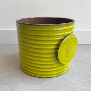 May include: A cylindrical, chartreuse-colored ceramic planter with a textured, ribbed design. A circular, decorative accent is attached to the side. The interior is a dark brown color. This decorative planter is suitable for indoor or outdoor use.