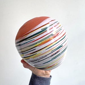 Könnte beinhalten: Eine Keramikschale mit runder Form, mit einer terrakottafarbenen Oberseite und einem weißen Boden. Die Schale ist mit farbenfrohen, diagonalen Pinselstrichen in Rot, Grün, Gelb und Schwarz verziert. Die Schale wird in der Hand einer Person gehalten.
