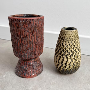 Puede incluir: Dos jarrones de cerámica. Uno es un jarrón alto, de color marrón rojizo, con una superficie texturizada similar a la corteza. El otro es un jarrón más pequeño, en forma de lágrima, con un patrón geométrico en tonos beige y negro. Ambos tienen interiores oscuros.