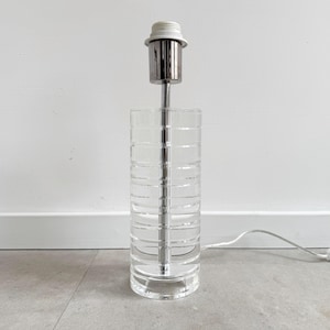 Könnte beinhalten: Eine Tischlampe aus klarem Glas mit einem silbernen Metallfuß und einem weißen Lampenschirm. Die Lampe hat ein strukturiertes, geripptes Design.