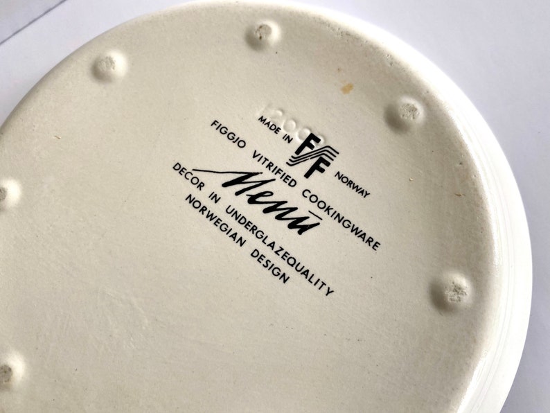 Pu&ograve; includere: Piatto in ceramica bianca con testo nero che recita "Made in Norway, Figgjo Vitrified Cookingware, Decor in Underglazequality Norwegian Design, Menu, FF".