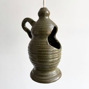 May include: A hanging, olive-green ceramic vessel with a handle and a large opening. The vessel has a bulbous shape with a textured, ridged surface. It is suspended by a natural-colored rope.