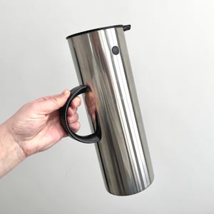 May include: A tall, stainless steel insulated carafe with a black handle and lid. The carafe has a cylindrical shape and a button on the side. The handle is curved and attached to the side of the carafe.