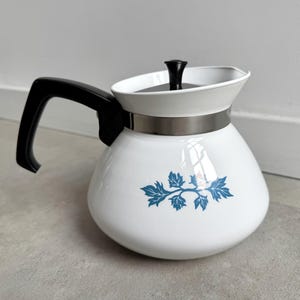 May include: White ceramic coffee pot with a black handle and lid. The pot features a blue floral design and a silver band around the middle. The handle is curved, and the lid has a black knob. A kitchenware item.