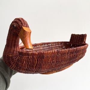 May include: A brown wicker duck-shaped basket with a wooden beak. The basket is made of woven wicker and has a detailed duck head and a small handle at the back. This decorative basket is suitable for storage or display.