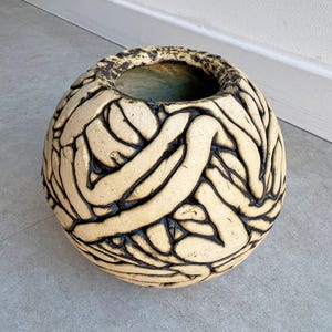 Pode incluir: Um vaso redondo de cerâmica com um design texturizado e tecido em preto e creme. O vaso tem uma abertura larga na parte superior.