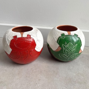 May include: Two spherical ceramic vases. One is red and white, the other is green and white. Both have a textured design resembling overalls. The vases have a brown interior and are suitable for decorative use.