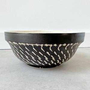 May include: A black ceramic bowl with a white, hand-painted design of diagonal lines. The bowl is slightly flared at the top and has a rounded bottom.