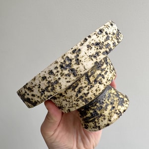 Puede incluir: Un cuenco de cerámica con pedestal y diseño moteado. El cuenco tiene una base de color crema con motas verdes oscuras y negras. El cuenco se sostiene en ángulo, mostrando su superficie texturizada y su forma única.