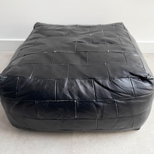 May include: A rectangular floor cushion constructed from multiple black leather panels stitched together in a brick-like pattern. The surface features a smooth, slightly reflective texture with visible seams connecting each individual rectangular piece.