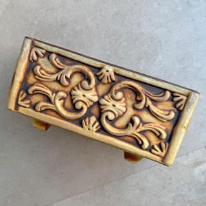 May include: A rectangular ceramic planter featuring a raised, ornate scrollwork pattern in shades of brown and cream. The piece has a weathered, antique finish with visible texture and is supported by small, integrated feet at the base.