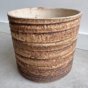 Pode incluir: Um vaso cilíndrico de cerâmica com um exterior texturizado e canelado num gradiente de tons terra castanhos e bege. O interior do vaso é de uma cor esbranquiçada lisa e o rebordo tem um acabamento orgânico ligeiramente irregular.