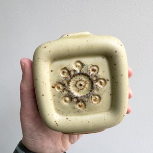 May include: A square, pale yellow ceramic vase with a raised floral design. The vase has a speckled texture and a small opening. The floral design is brown and circular. The vase is held in a hand.