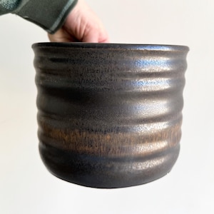 May include: A handmade, dark brown ceramic planter with a textured, ridged design. The planter has a metallic sheen and a wide, cylindrical shape. The interior is not visible. The planter is held by a person's hand.