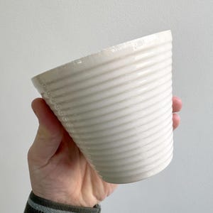 Pode incluir: Um vaso de cerâmica branco com um design canelado. O vaso é segurado em uma mão e está inclinado para o espectador. O vaso tem uma estética simples e moderna.