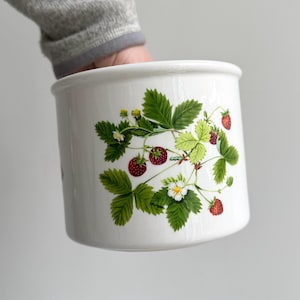 Puede incluir: Contenedor de cerámica blanca con diseño de planta de fresa. El diseño presenta hojas verdes, flores blancas y fresas rojas maduras. El contenedor es cilíndrico con un borde ligeramente ensanchado.